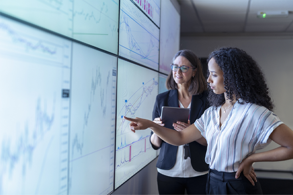 One woman pointing at giant screen and another woman with clipboard next to her