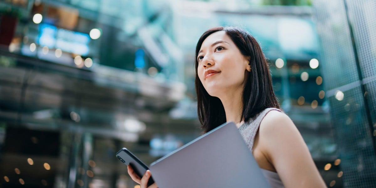 a finance leader smiles confidently while holding a mobile phone in one hand and a laptop under her other arm