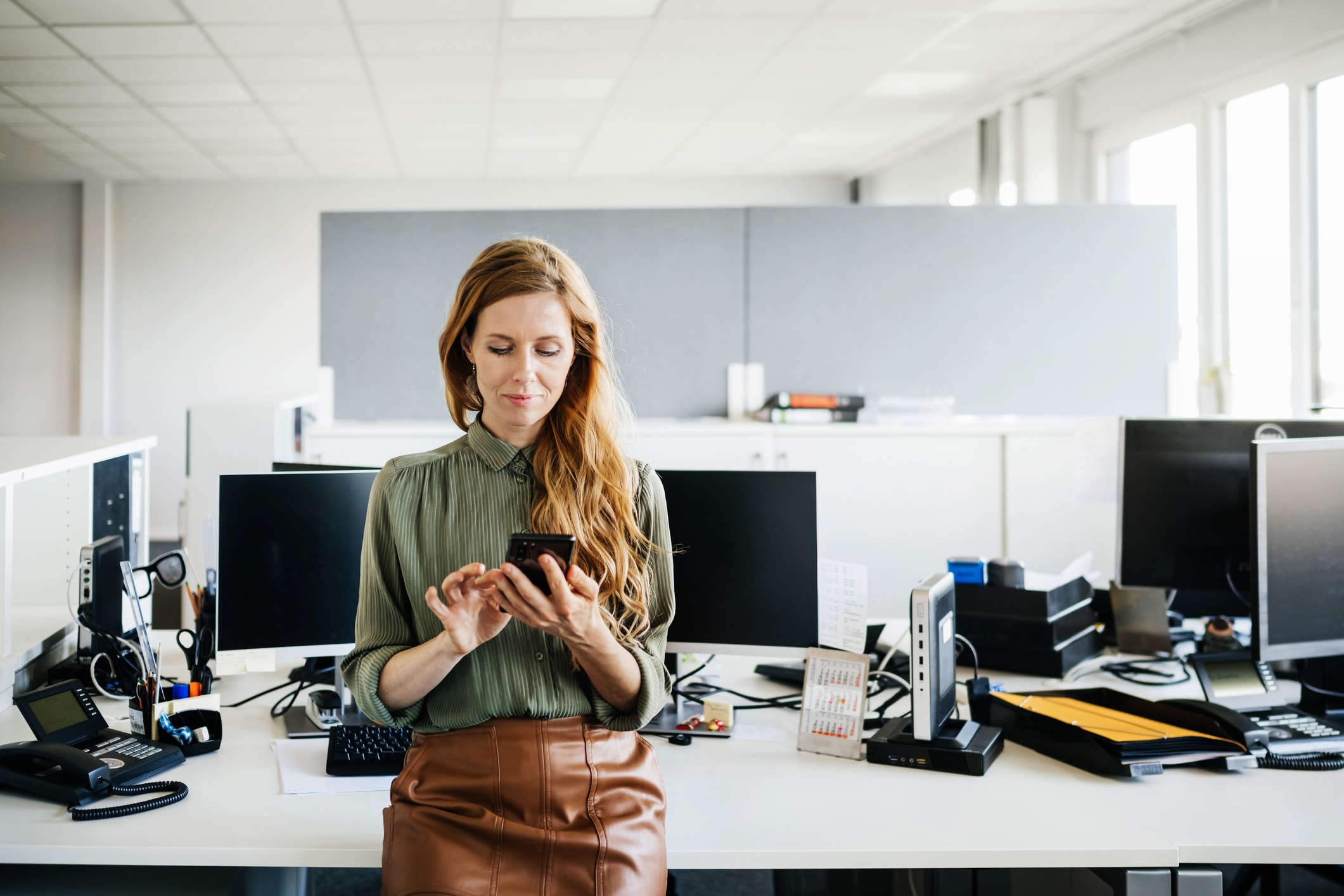 Woman leaning against a desk of computers looking at her phone