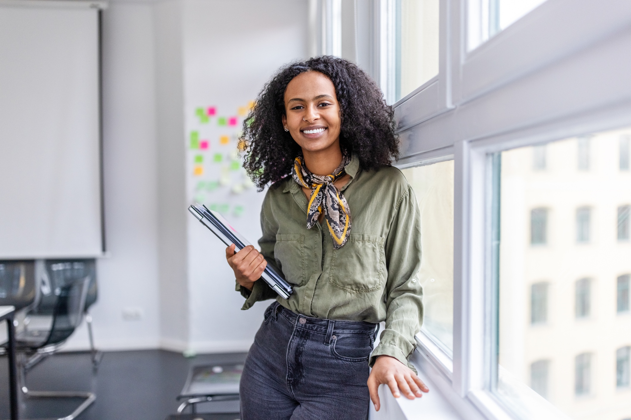 Woman leaning against a window with a closed laptop in her arms smiling at the camera