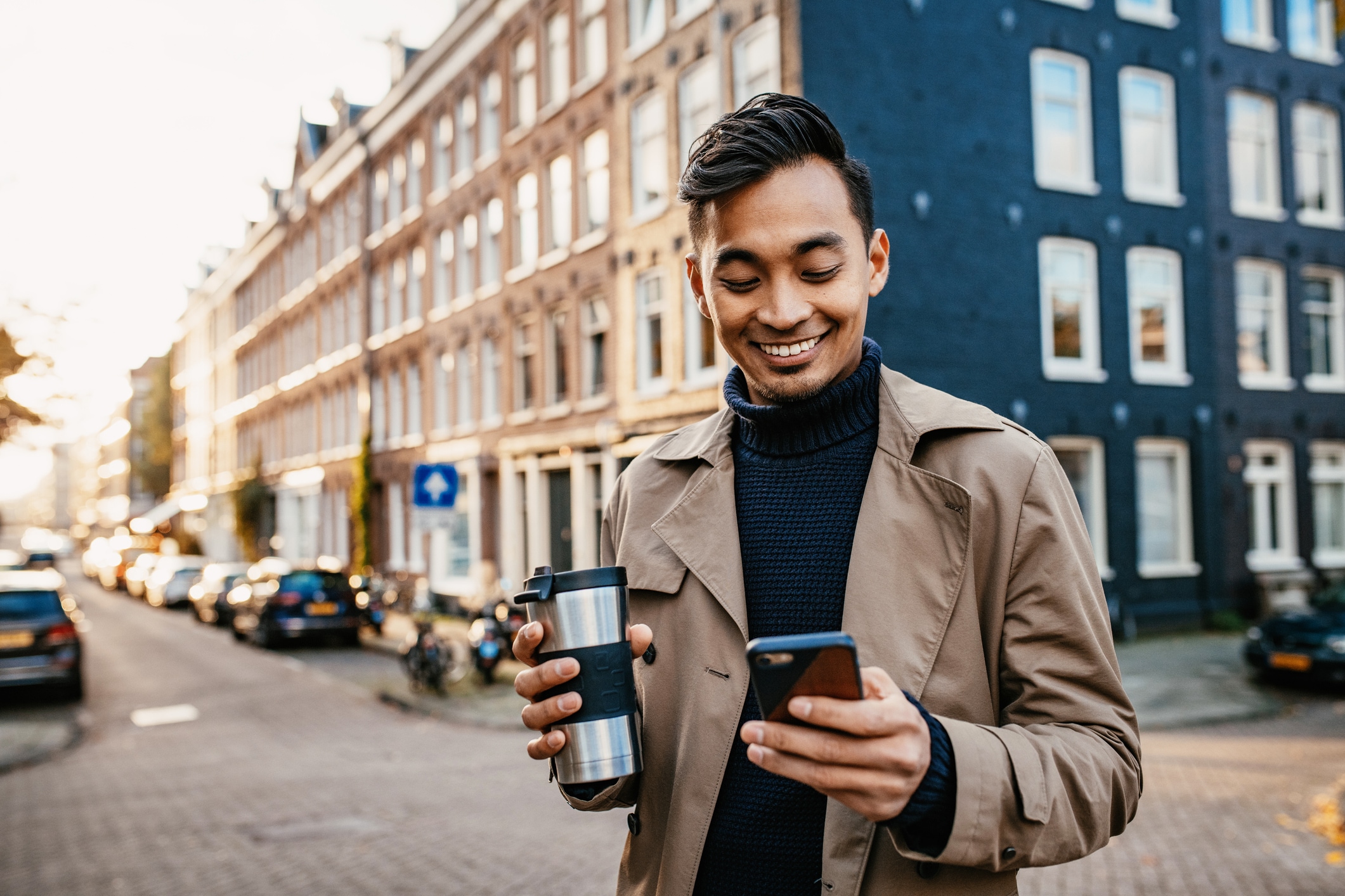 Man looking at phone outside with a coffee tumbler in his hand smiling