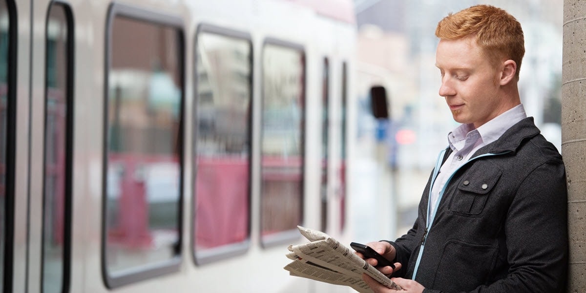 man standing next to train holding newspaper and cell phone