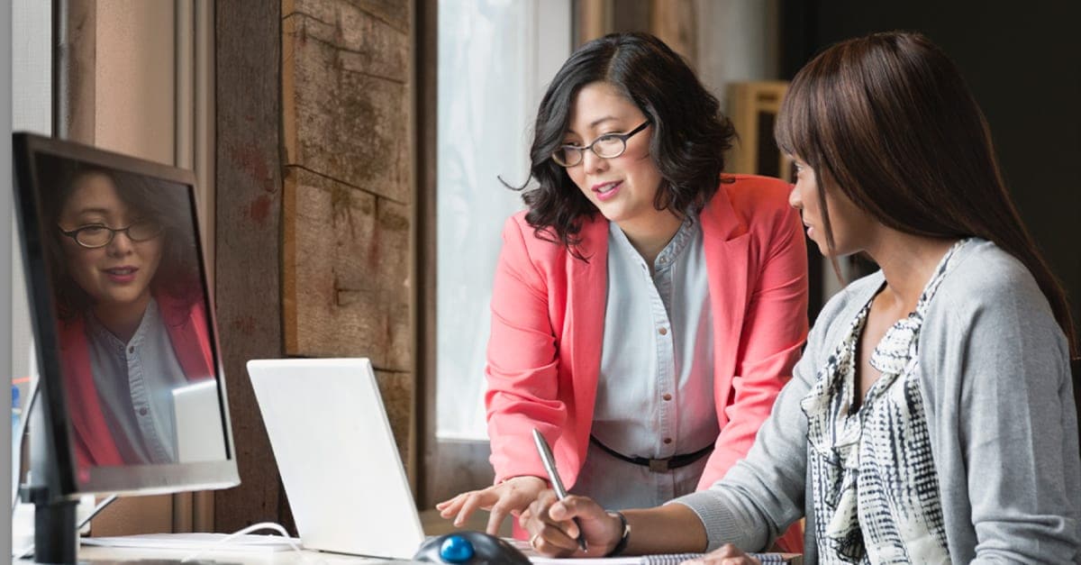 Woman seated at an office computer desk collaborates with another woman standing next to her