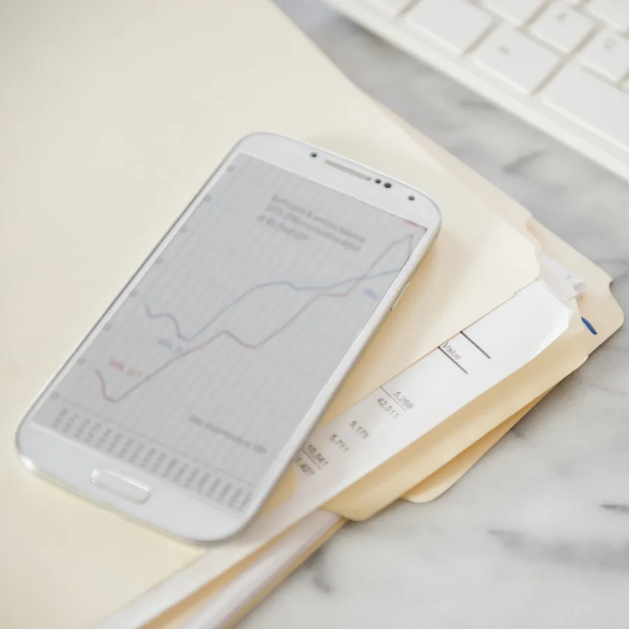 A smartphone displaying a financial line chart rests on top of printed financial documents on a marble desk, with a white keyboard and glasses in the background.