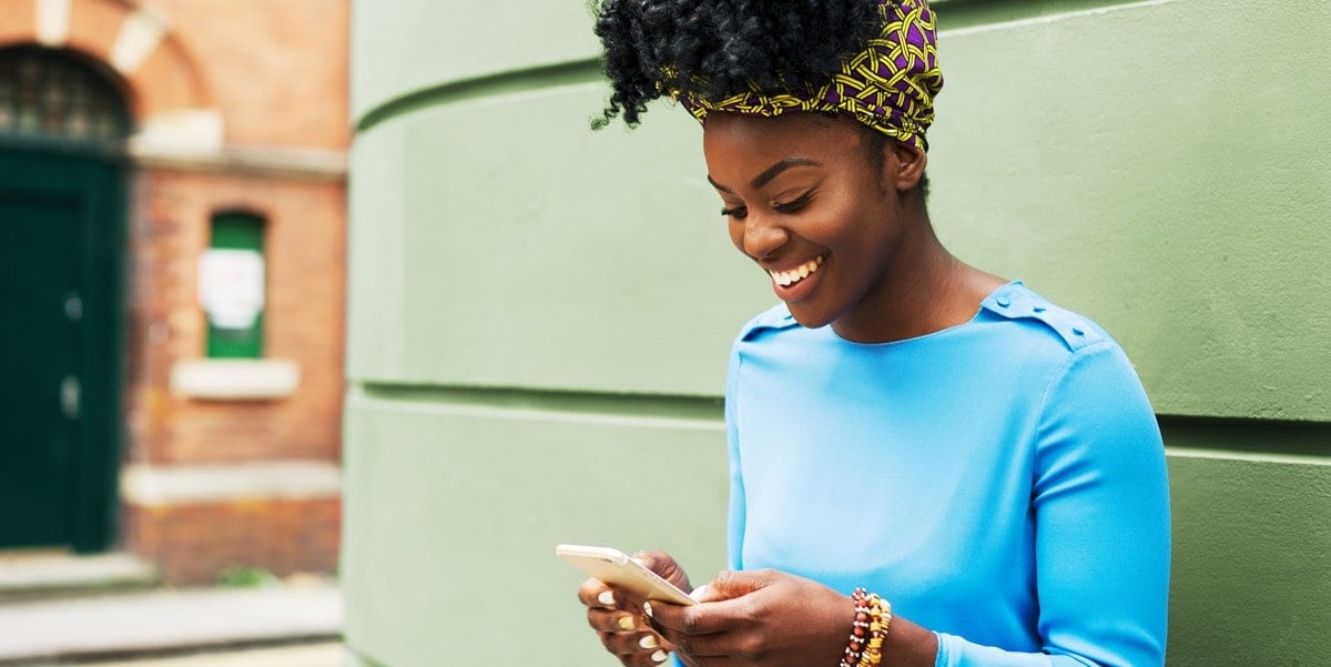 Woman wearing blue top stands by green wall, looking down at cell phone smiling