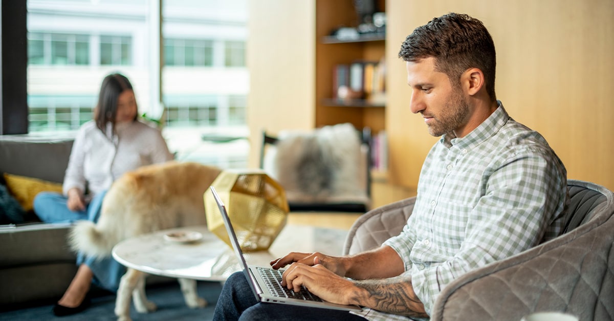 Man working from home on a laptop, while a woman pets a dog in the background
