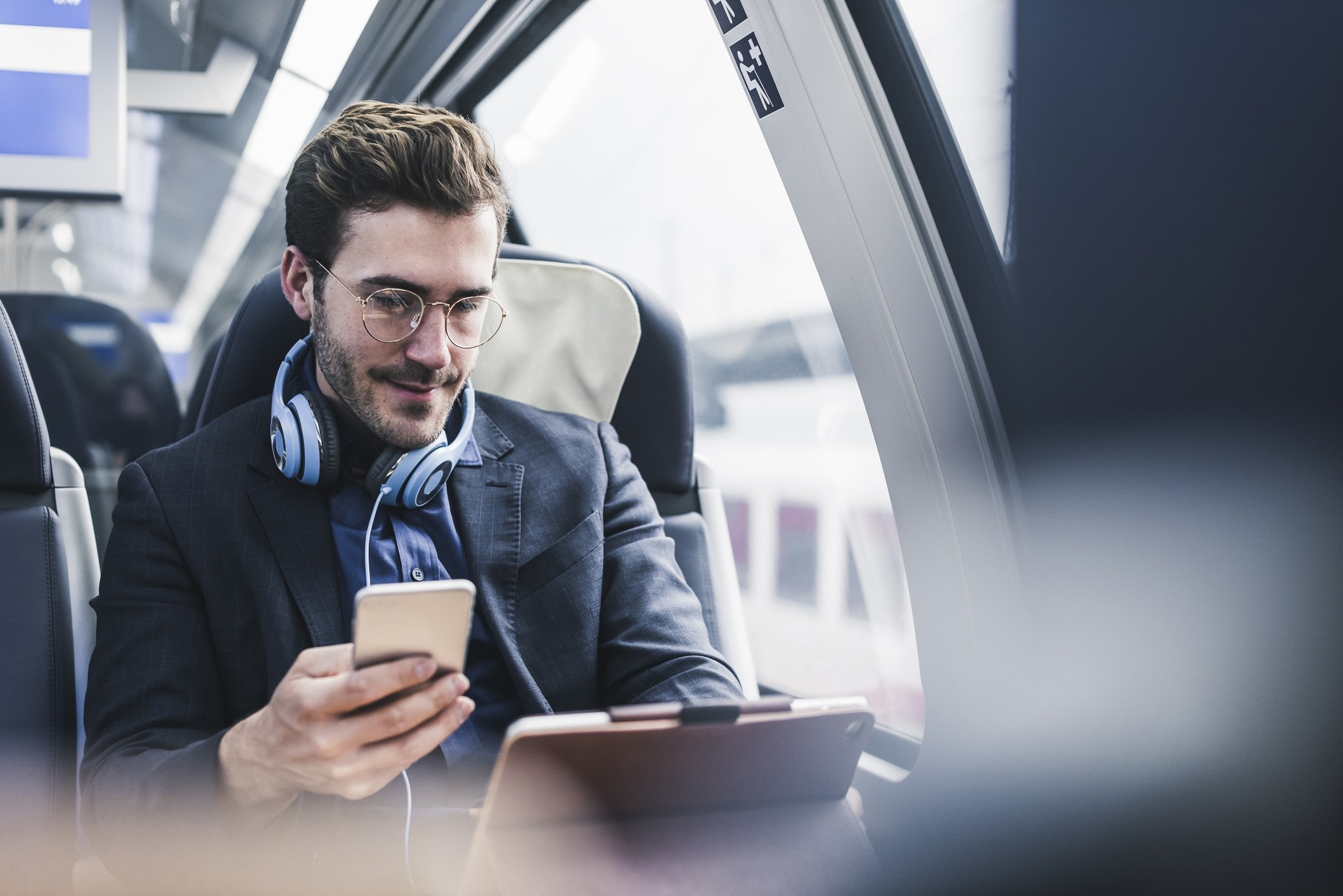 Man with glasses sitting on a train, wearing glasses, holding phone, looking at tablet 